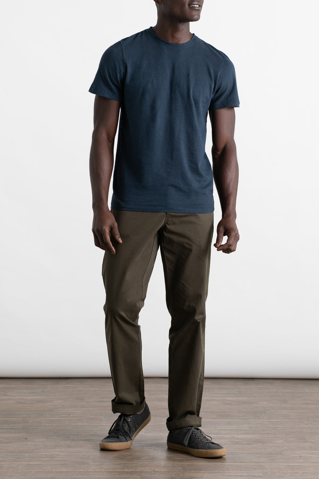 A person stands indoors on a wooden floor in front of a plain, light background, wearing a Jungmaven Baja Tee in Navy and olive green pants. Their face is not visible. They also wear black sneakers with tan soles.