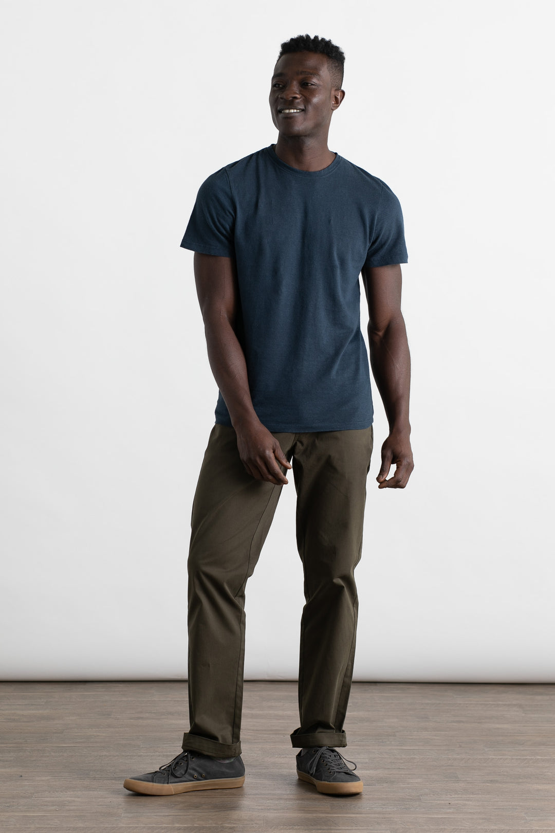 A man stands indoors against a plain white background, smiling. He is wearing the Jungmaven Baja Tee in Navy by Jungmaven, paired with olive green pants and gray sneakers. The floor is wooden, and his hands are relaxed by his sides.