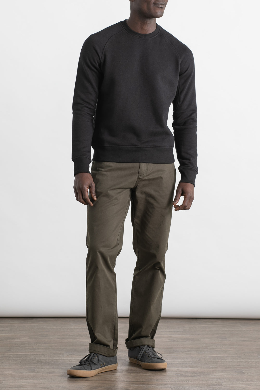 A man stands indoors against a plain white background, wearing a Bridge & Burn Fremont Crew Sweatshirt in black, olive green pants, and gray sneakers. His face is not visible.