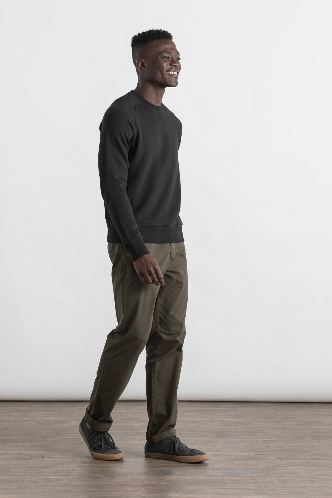 A man smiles while walking indoors against a plain white background, wearing the Bridge & Burn Fremont Crew Sweatshirt in black, olive green pants, and black sneakers.