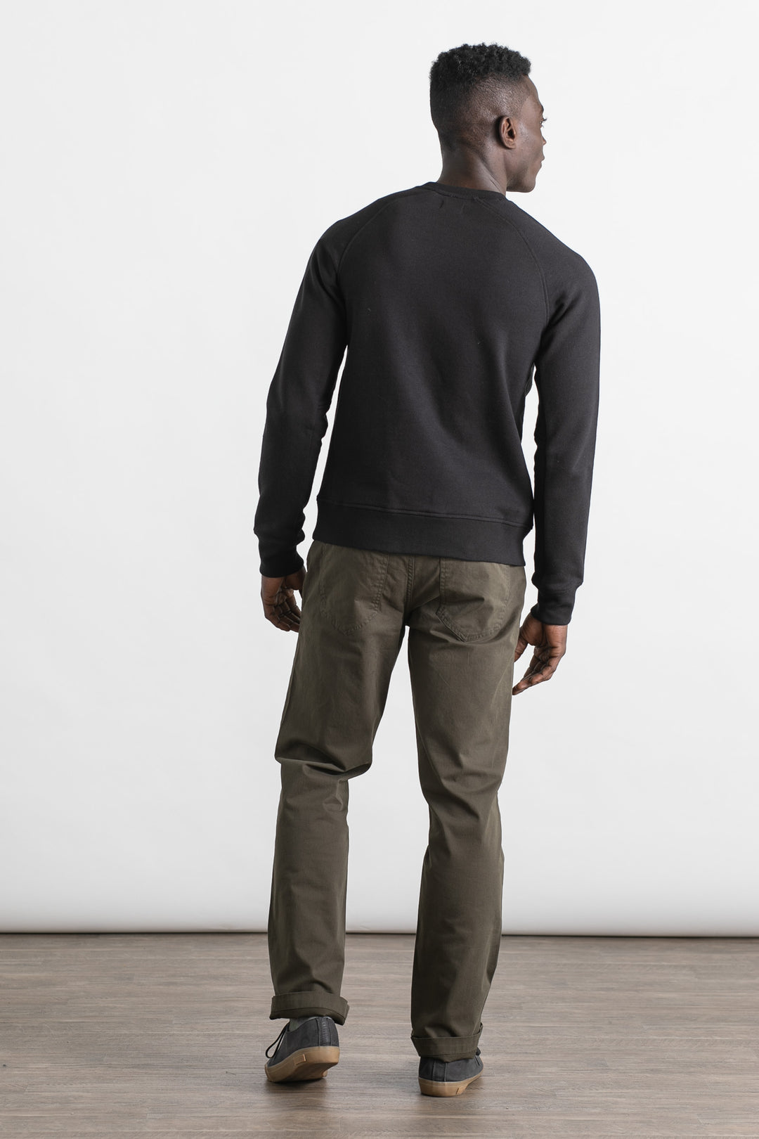 A man stands indoors on a wooden floor facing away from the camera, wearing the Bridge & Burn Fremont Crew Sweatshirt in black, olive green pants, and black sneakers. The background is plain white.