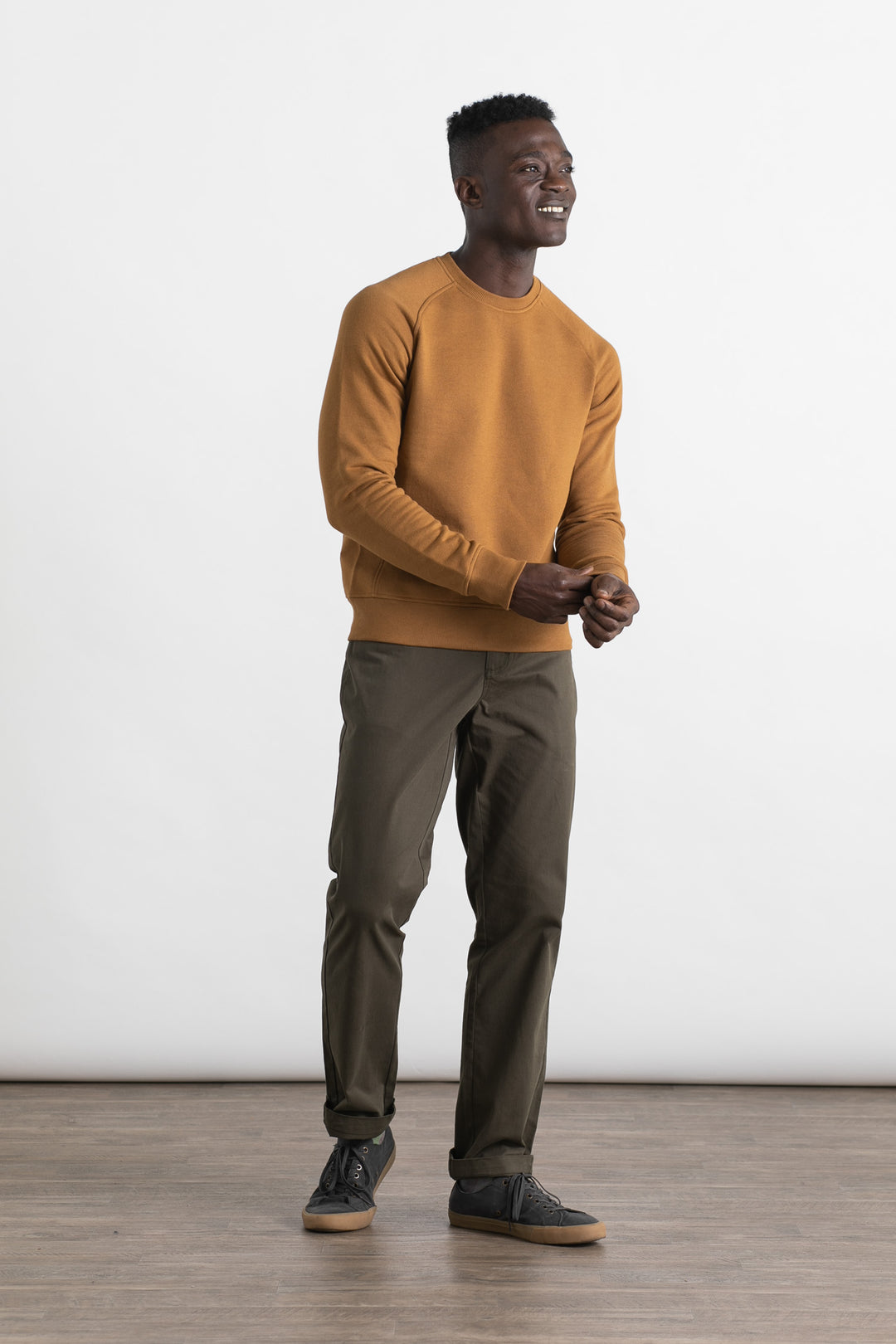 A man smiles while wearing the Bridge & Burn Fremont Crew Sweatshirt in bronze, paired with olive green pants and black sneakers with gum soles. He stands against a plain white background on a wooden floor.