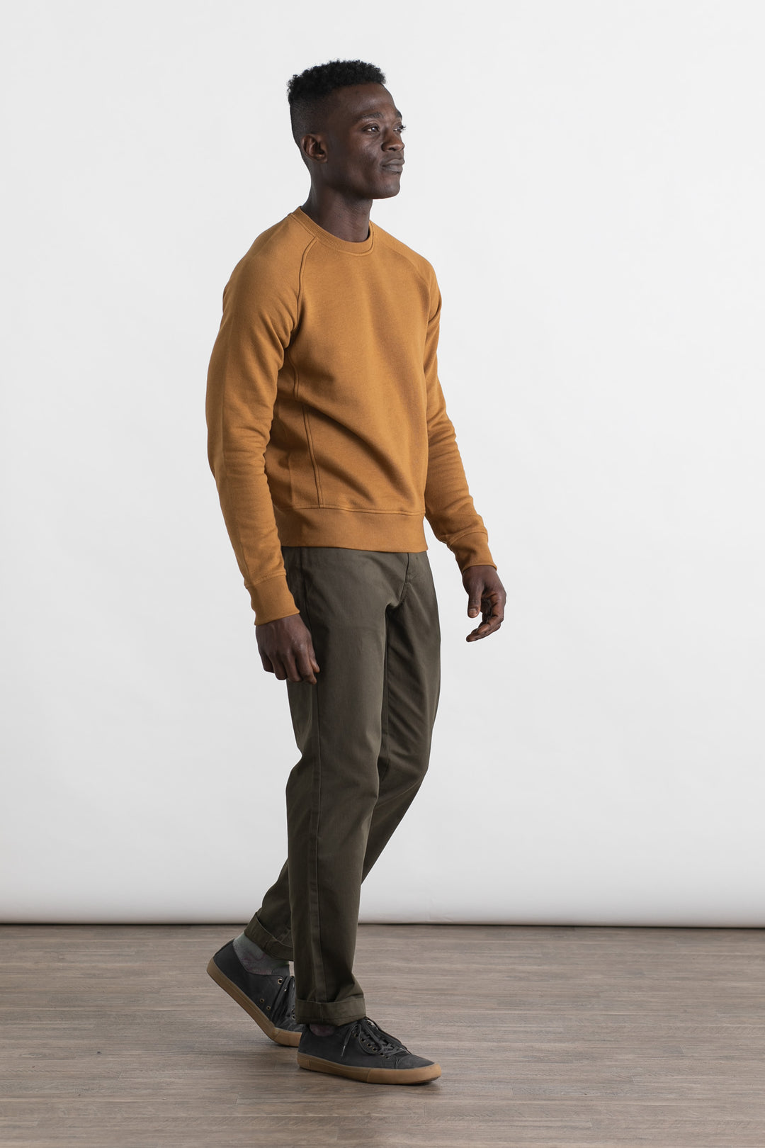 A man wears the Bridge & Burn Fremont Crew Sweatshirt in bronze, made from organic cotton, styled with olive green pants and dark sneakers, walking on a wooden floor against a plain white background.