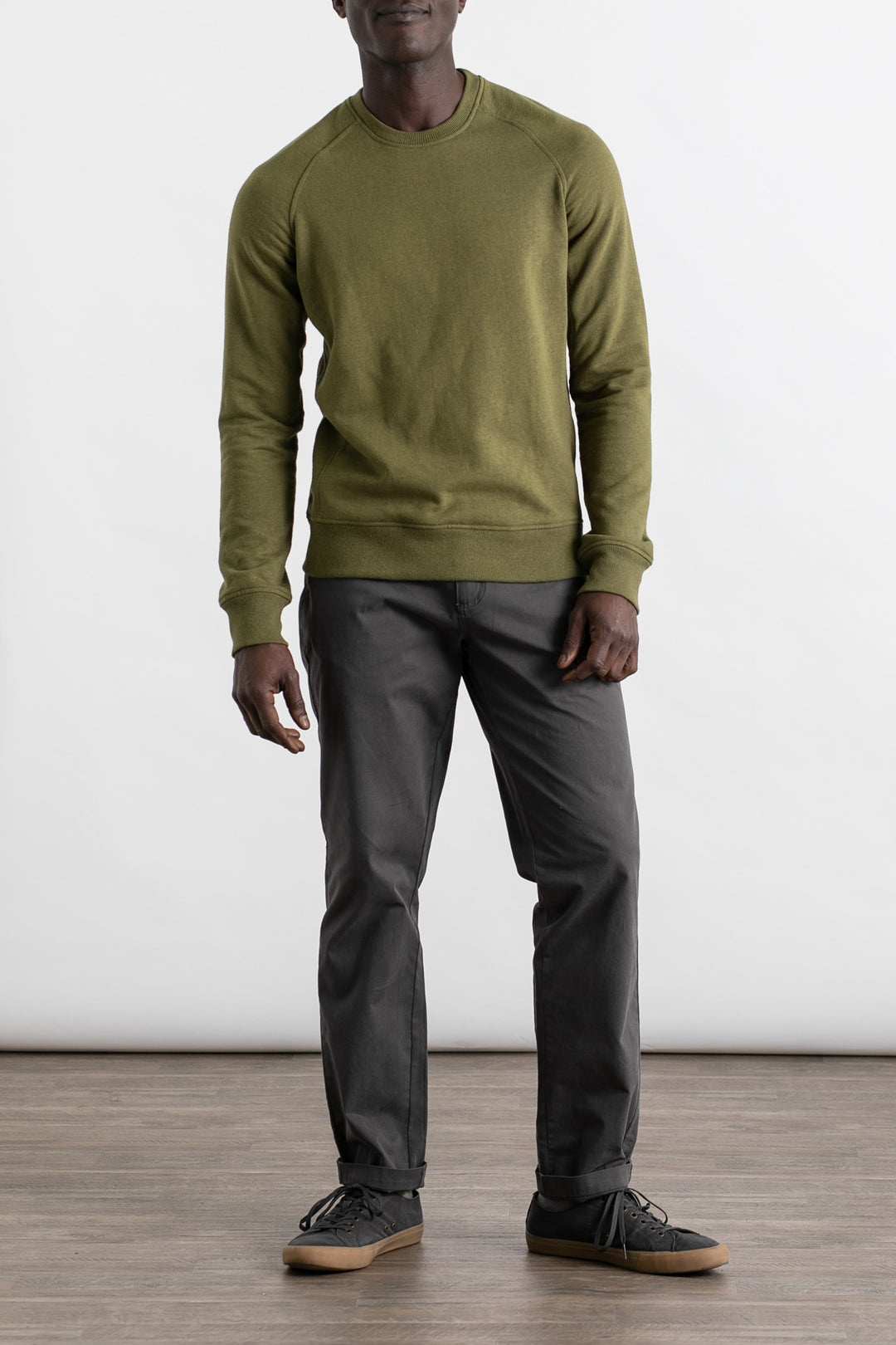 A person wearing the Bridge & Burn Fremont Crew Sweatshirt in olive, along with gray pants and black sneakers, stands indoors on a wooden floor against a plain white background. Their face is partially obscured.