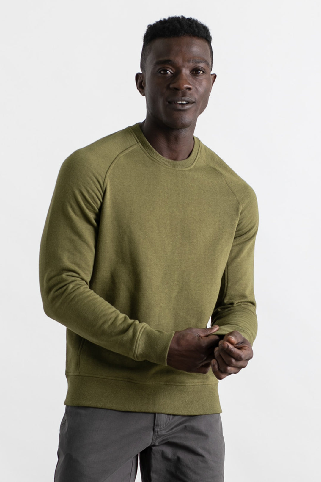 A man wearing the Bridge & Burn Fremont Crew Sweatshirt in olive and gray pants stands against a plain white background, holding one hand with the other and looking forward with a slight smile.