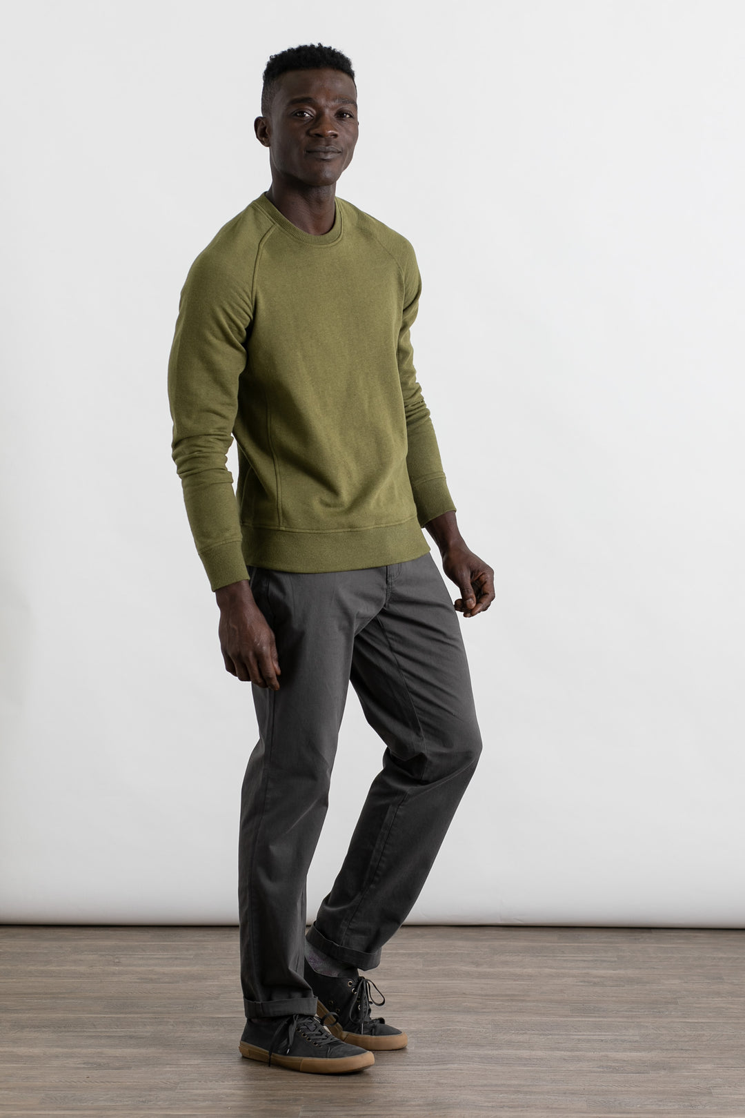 A man stands on a wooden floor against a white background, wearing the Bridge & Burn Fremont Crew Sweatshirt in olive, gray pants, and gray sneakers. He is smiling slightly and facing the camera.