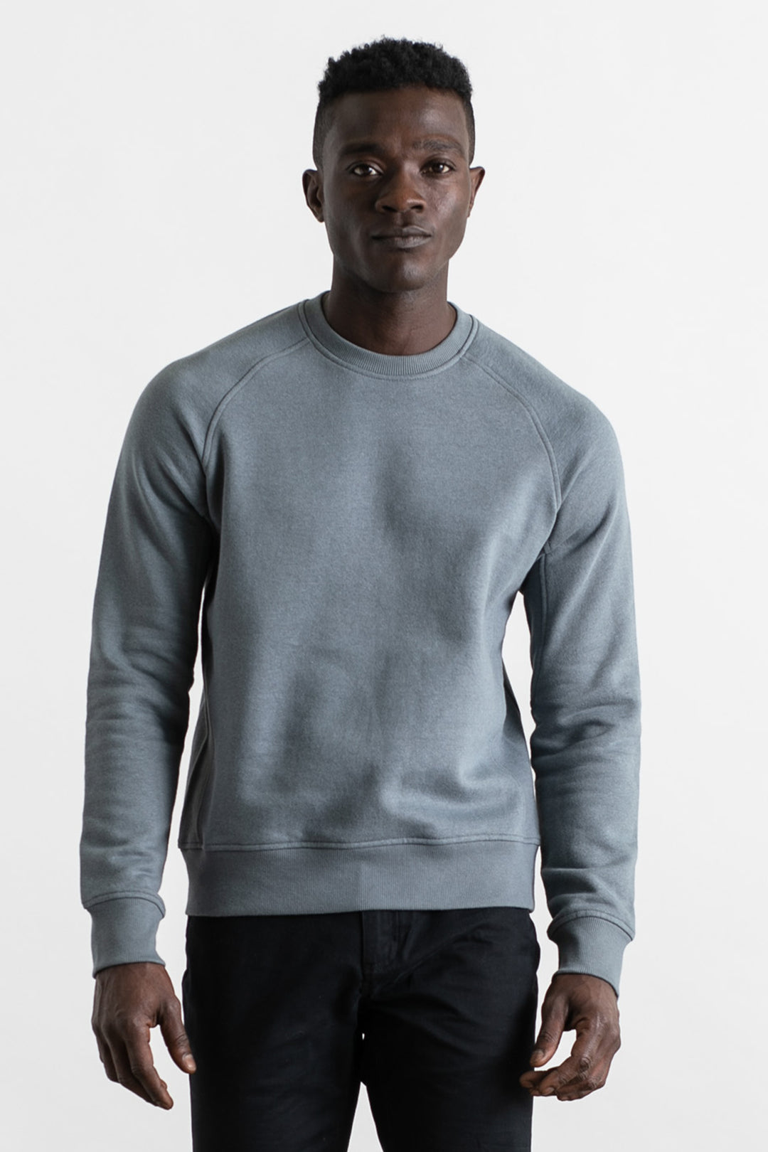 A man wears the Bridge & Burn Fremont Crew Sweatshirt in Storm Blue and black pants, standing facing the camera against a plain white background.