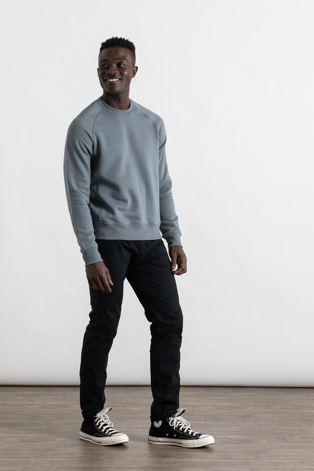 A man wears the Bridge & Burn Fremont Crew Sweatshirt in Storm Blue, paired with black pants and high-top sneakers, standing on a wooden floor against a plain white background, smiling and glancing to the side.