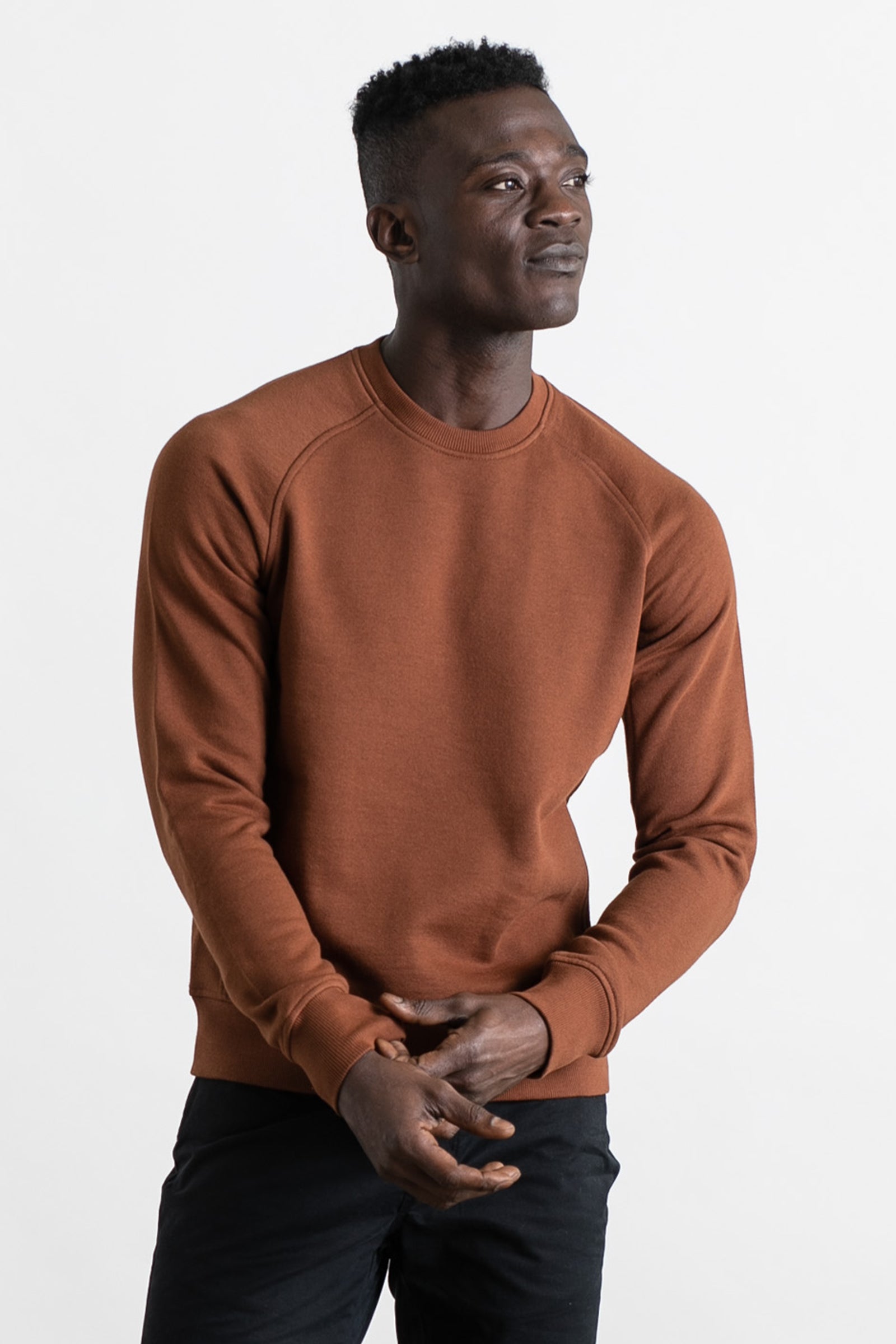 A man in the Bridge & Burn Fremont Crew Sweatshirt in Burnt Umber and black pants stands against a white background, gazing to the side with a slight smile and hands loosely clasped.