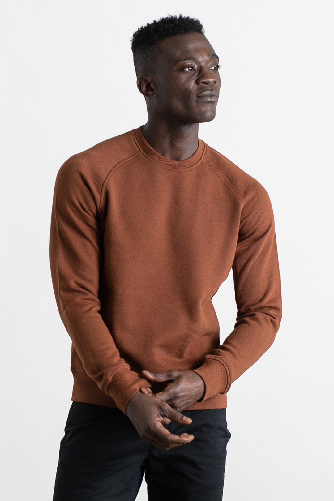 A man in the Bridge & Burn Fremont Crew Sweatshirt in Burnt Umber and black pants stands against a white background, gazing to the side with a slight smile and hands loosely clasped.