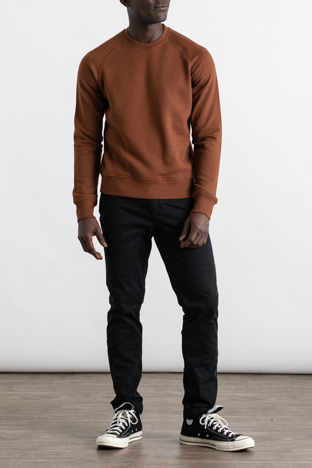 A person stands indoors wearing the Bridge & Burn Fremont Crew Sweatshirt in Burnt Umber, paired with black pants and black high-top sneakers with white soles, against a plain white background and wooden floor.