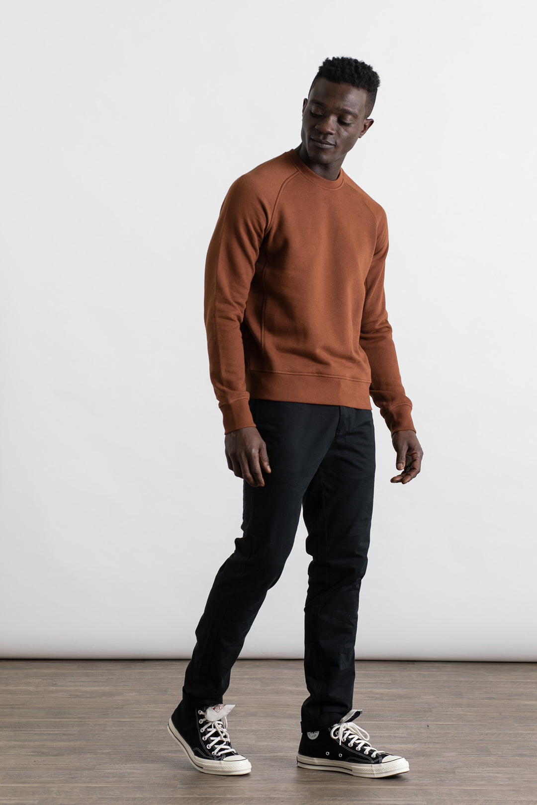A man stands indoors against a plain white background, wearing the Bridge & Burn Fremont Crew Sweatshirt in Burnt Umber, black pants, and black high-top sneakers. He looks slightly left with a relaxed stance.