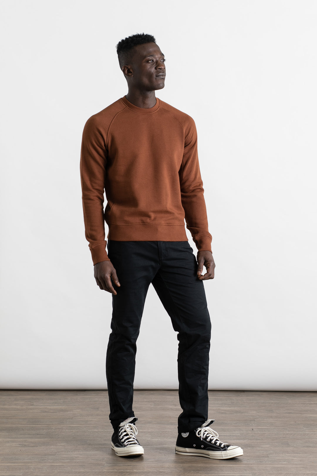 A man stands indoors on a wooden floor, wearing the Bridge & Burn Fremont Crew Sweatshirt in Burnt Umber, paired with black pants and black-and-white sneakers. He looks slightly to the side with a neutral expression against a plain white background.