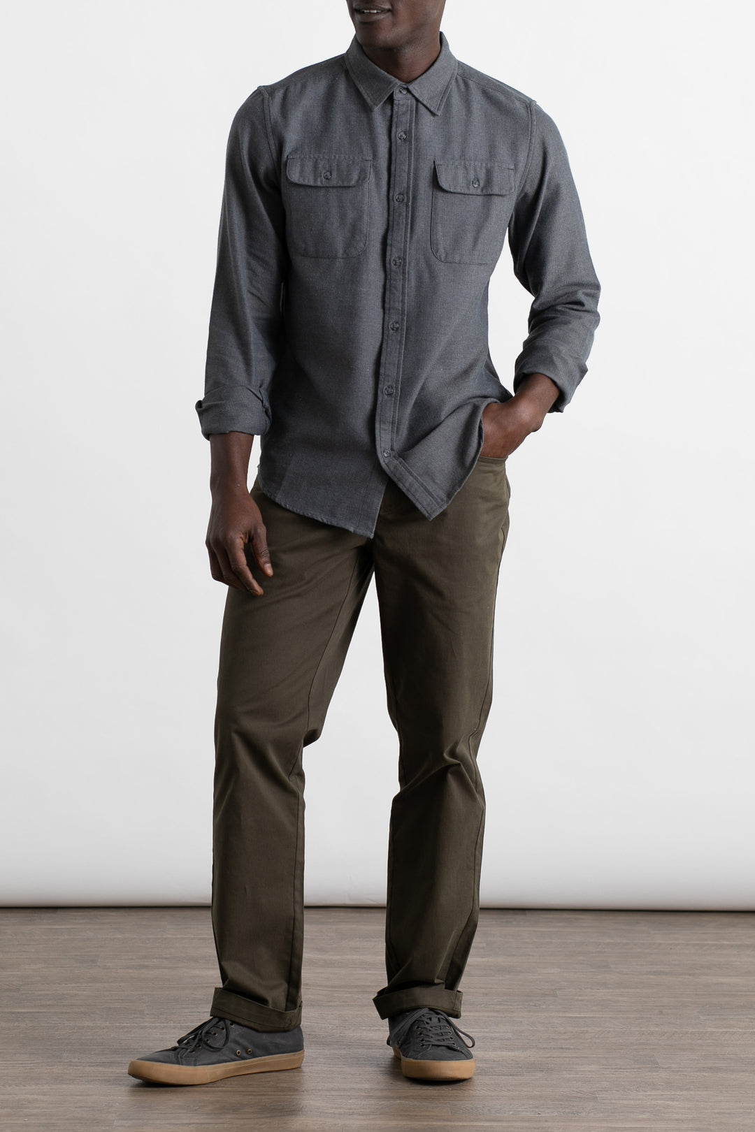 A man stands indoors against a plain white background wearing the Bridge & Burn Bedford Shirt in Charcoal Herringbone with chest pockets, paired with olive green pants and gray sneakers with tan soles. One hand is in his pocket.