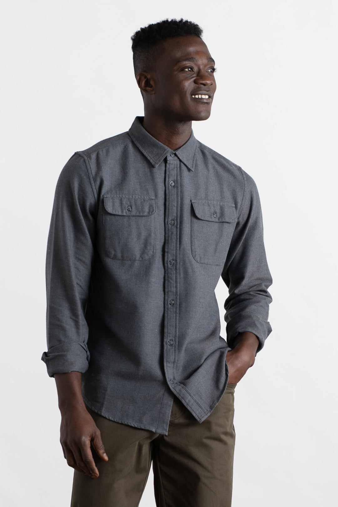 A man wearing the Bridge & Burn Bedford Shirt in Charcoal Herringbone with olive pants stands against a white background, smiling and looking to the side with one hand in his pocket.