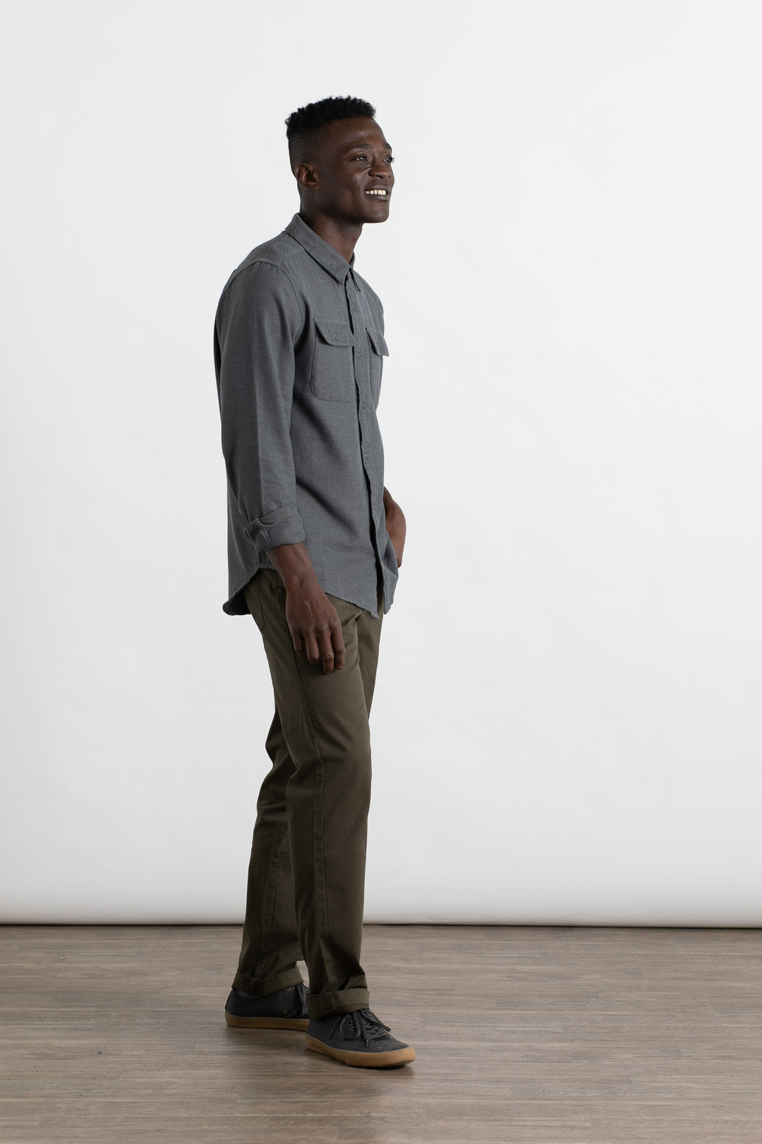 A man stands indoors on a wooden floor by a plain white background, smiling. He is wearing the Bridge & Burn Bedford Shirt in Charcoal Herringbone, olive green pants, and dark sneakers.