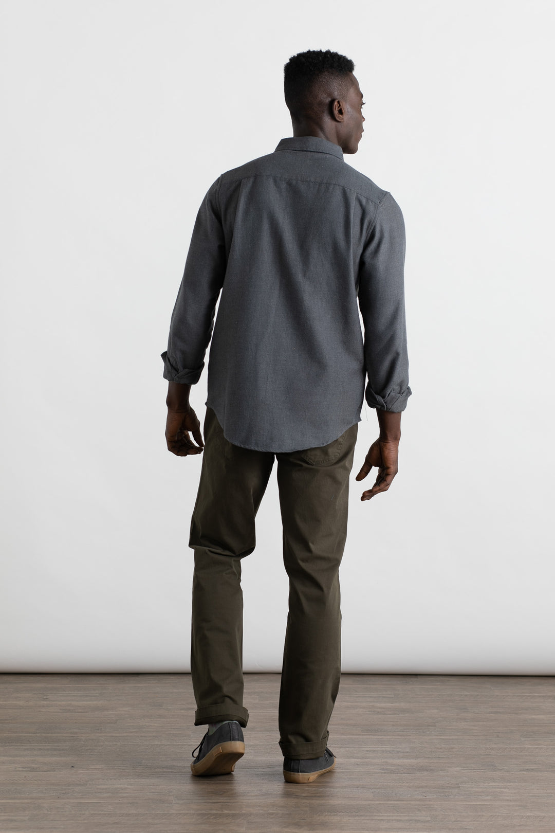 A man stands indoors on a wooden floor facing away from the camera, wearing the Bridge & Burn Bedford Shirt in Charcoal Herringbone, olive green pants, and gray shoes. The background is plain and white.
