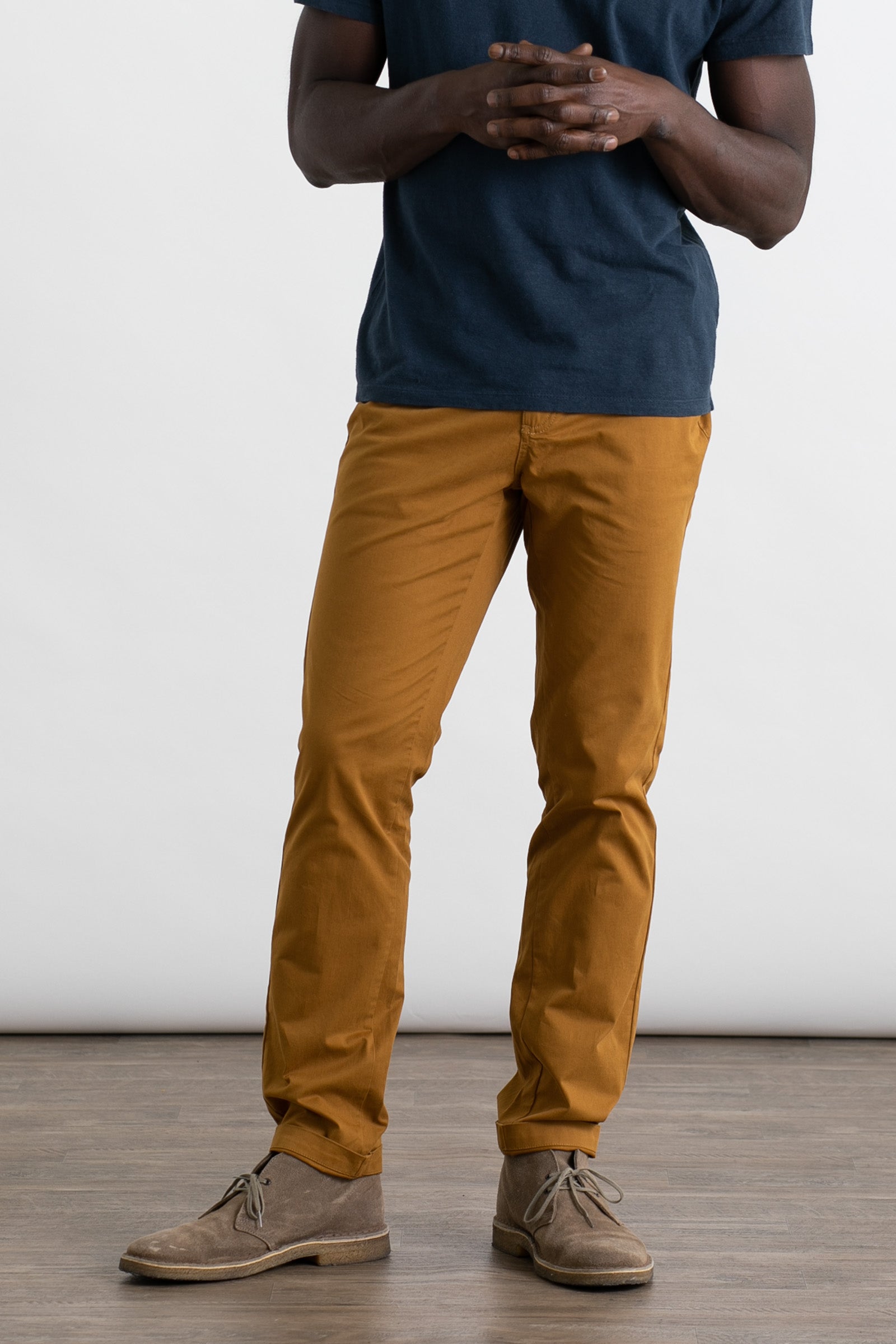 A person stands indoors on a wooden floor wearing Bridge & Burn's Bradley Slim Pant in Flax. They pair the pants with a dark blue t-shirt and beige suede shoes, hands clasped together; the image does not show their head.