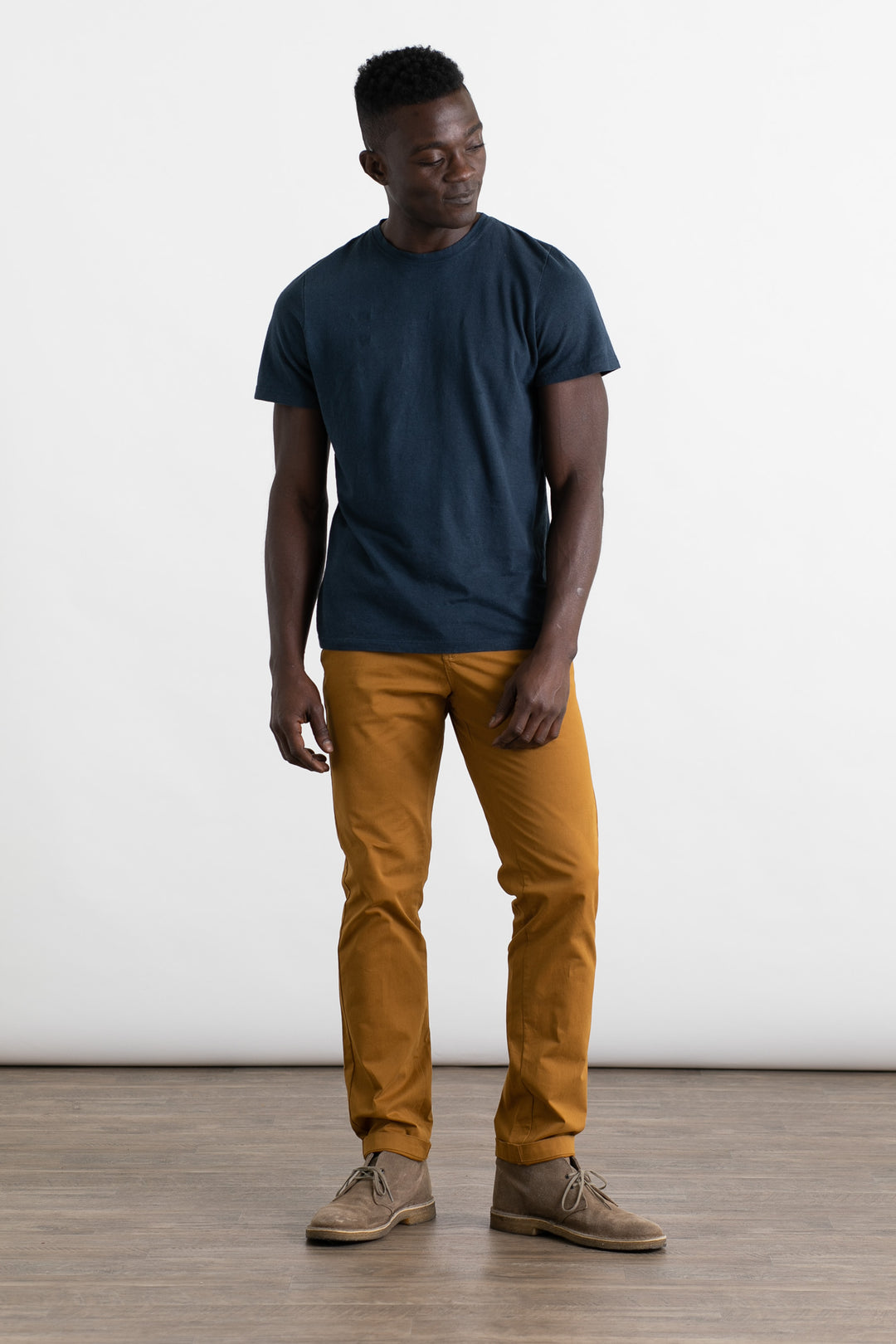 A man stands on a wooden floor against a white background, wearing a dark blue t-shirt and Bridge & Burn Bradley Slim Pant in Flax. The 5-pocket style of these pants adds a modern touch to his relaxed look.