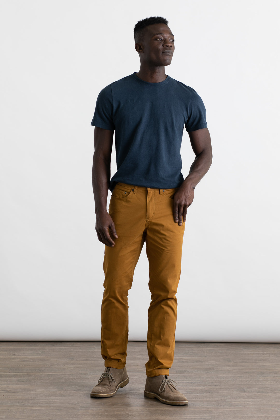 A man stands on a wooden floor against a white background, wearing a navy blue t-shirt, Bridge & Burn Bradley Slim Pant in Flax, and light brown shoes. He looks to the side with a relaxed expression and one hand in his pocket.