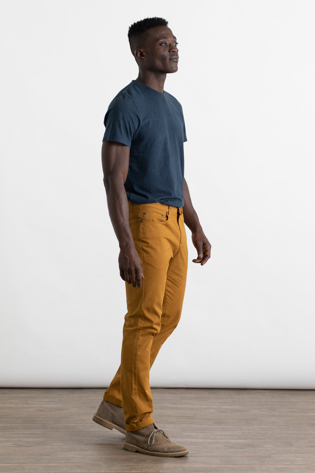 A man poses confidently in a studio, wearing a navy t-shirt and Bridge & Burn's Bradley Slim Pant in Flax, paired with light brown shoes. He stands against a plain white background on a wooden floor.