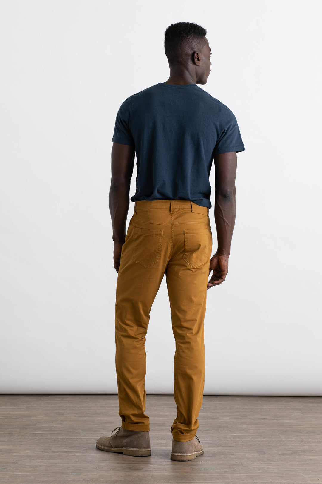 A man stands facing away, wearing a navy t-shirt and Bridge & Burn’s Bradley Slim Pant in Flax with matching shoes. He is on a wooden floor against a plain white background.