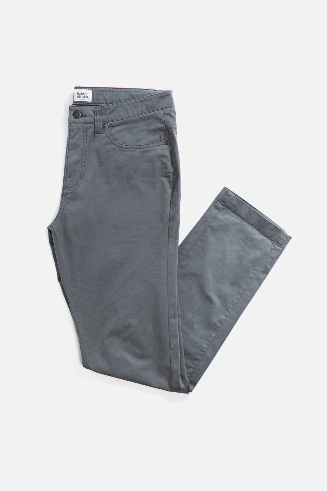 A pair of Bradley Slim Pant in Storm Blue by Bridge & Burn, neatly folded on a white background. These men's slim fit pants have 5 pockets and a visible brand label on the waistband.