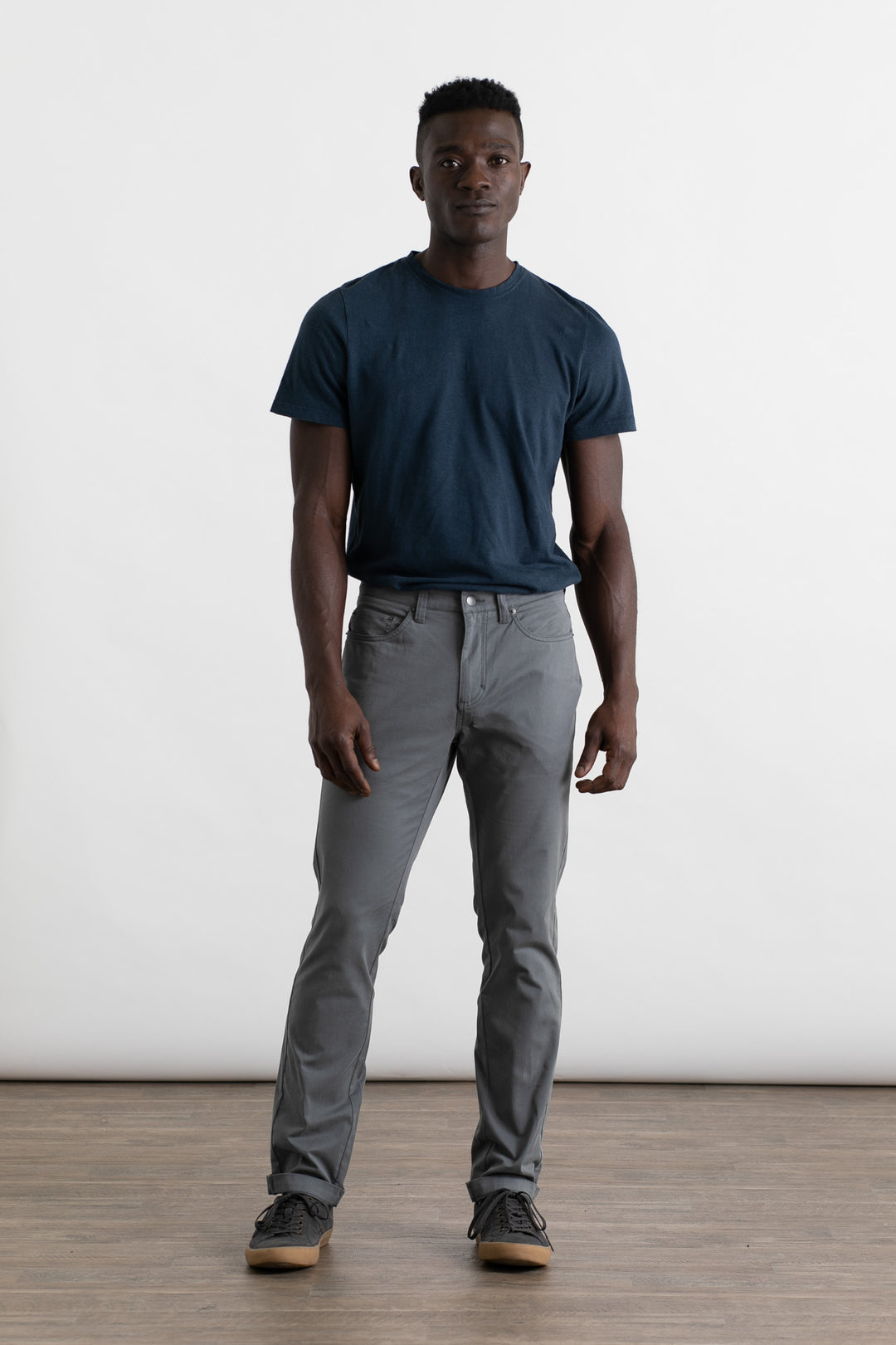 A man stands on a wooden floor against a plain white background, wearing a dark blue t-shirt and Bridge & Burn’s Bradley Slim Pant in Storm Blue, featuring a 5-pocket style. He faces forward with a neutral expression and relaxed posture.