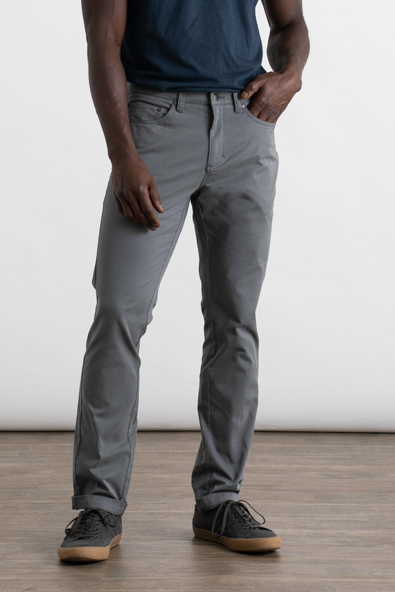A person in a navy blue t-shirt, Bridge & Burn's Bradley Slim Pant in Storm Blue, and dark sneakers stands on a wooden floor with one hand in their pocket against a plain light background.