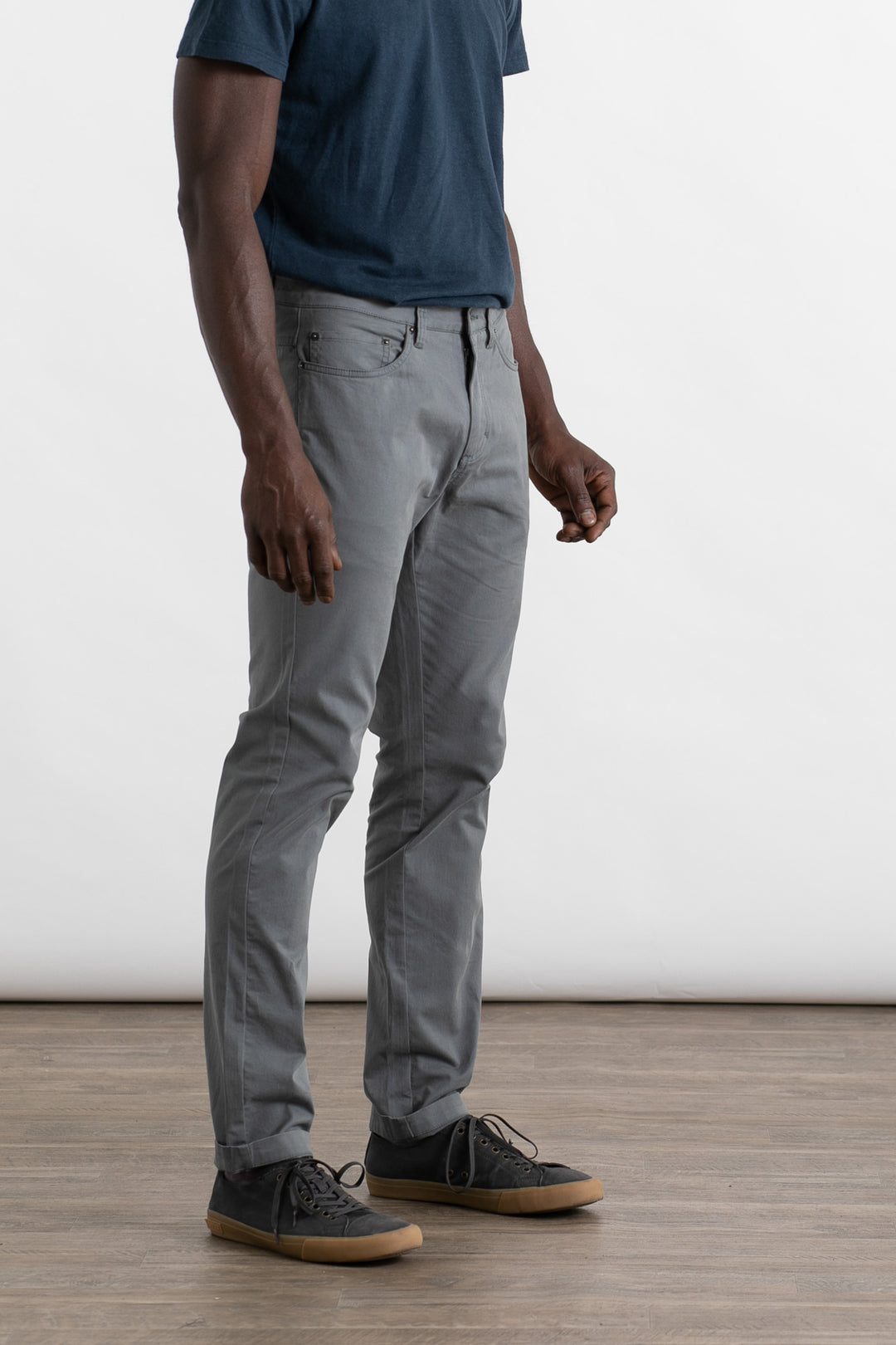 A person stands on a wooden floor against a plain white background, wearing Bridge & Burn's Bradley Slim Pant in Storm Blue. They also wear a dark blue t-shirt and black sneakers with brown soles; the upper part of their head is not visible.
