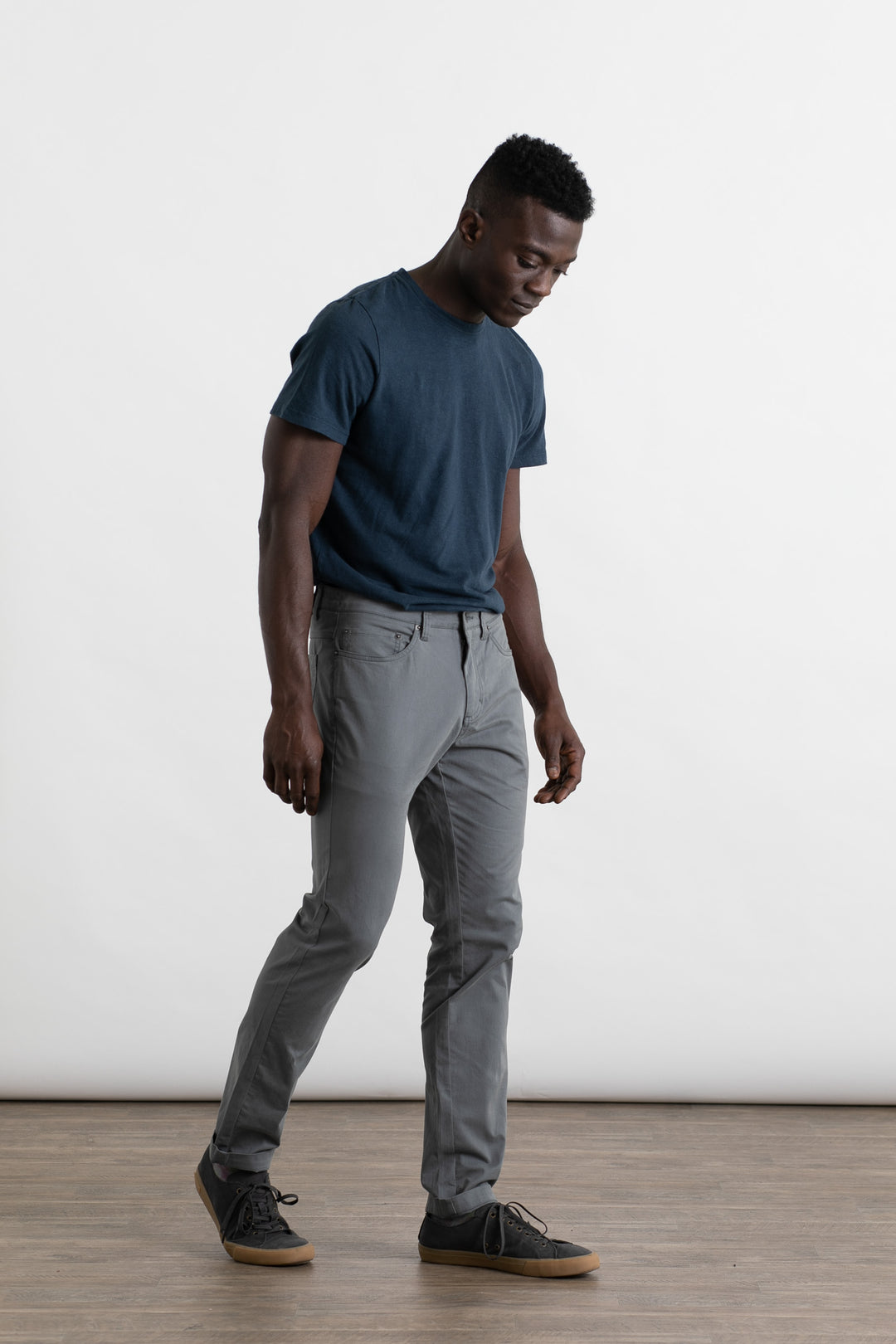 A man wearing Bridge & Burn's Bradley Slim Pant in Storm Blue walks indoors on a wooden floor against a plain white background, looking slightly downward.