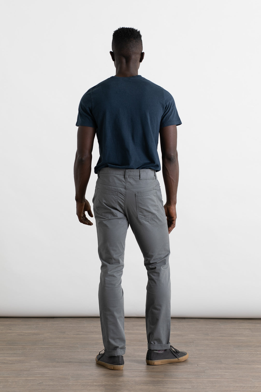 A man faces away from the camera wearing Bridge & Burn's Bradley Slim Pant in Storm Blue, a navy blue t-shirt, and dark sneakers. He stands on a wooden floor against a plain white background.