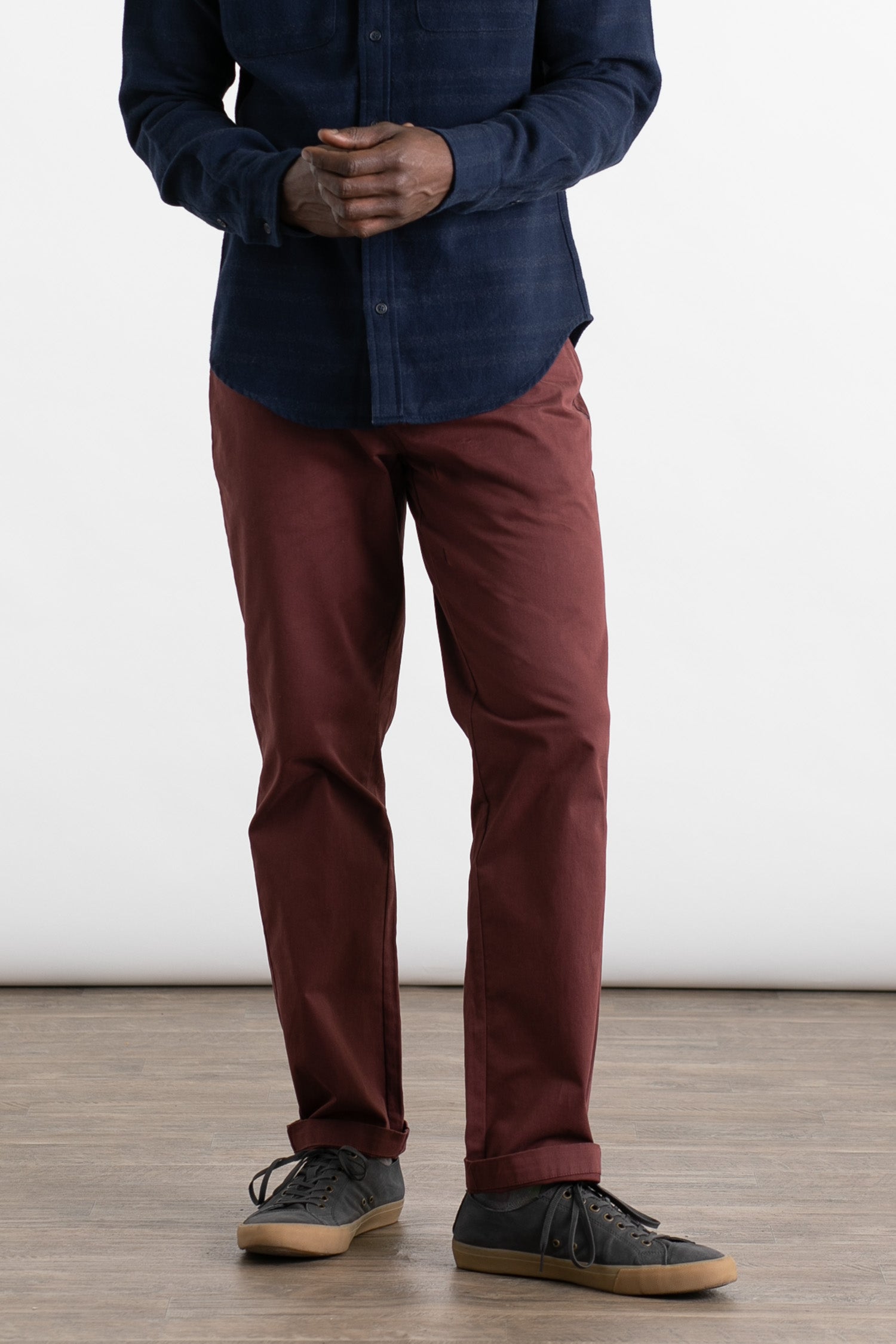 A person wears Bridge & Burn’s Polk Pant in Burgundy, a straight-fit, 5-pocket organic cotton style, paired with a dark blue button-up shirt and black sneakers with gum soles. They stand indoors on a wooden floor against a plain white background. Face not visible.