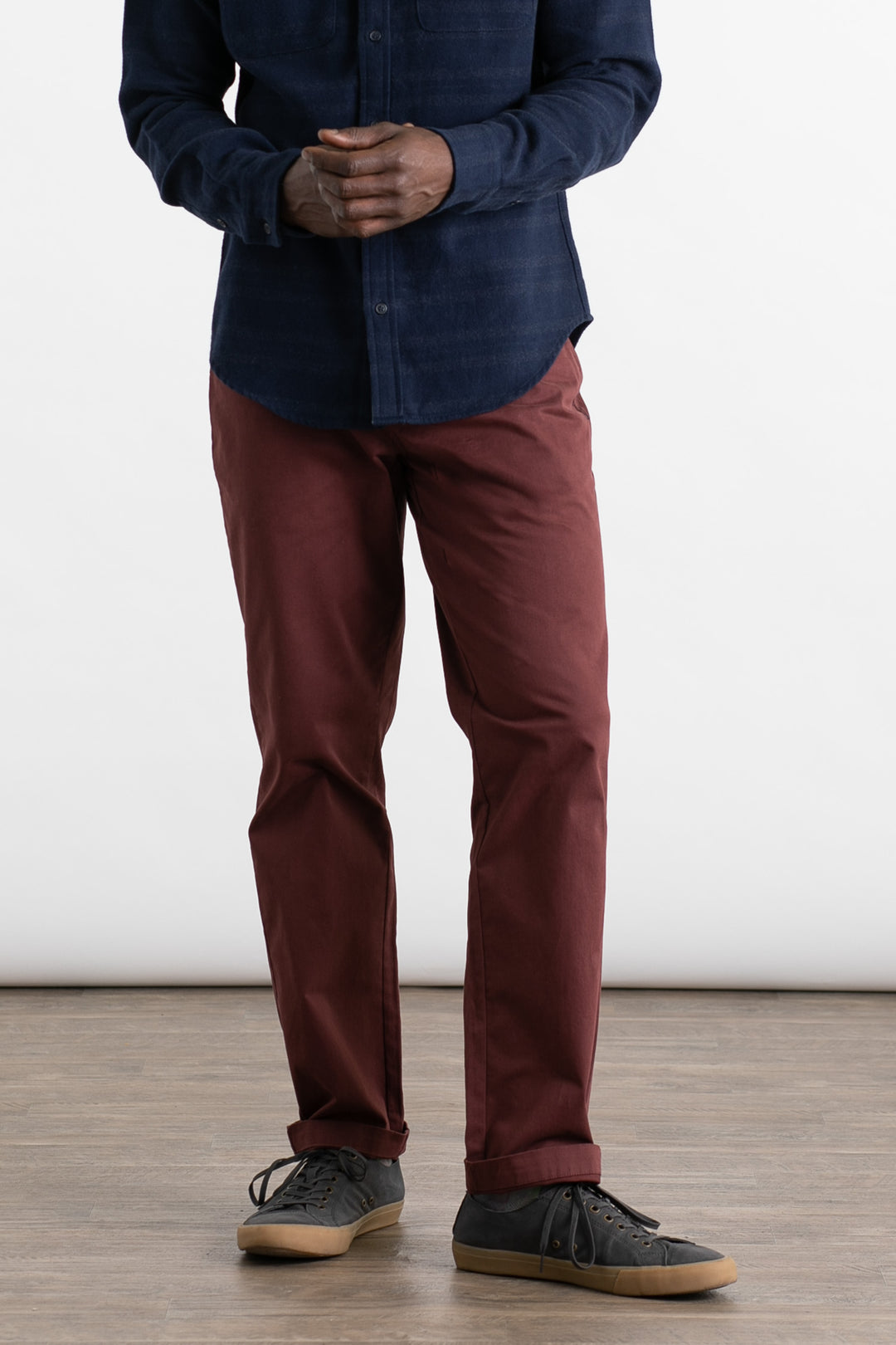 A person wears Bridge & Burn’s Polk Pant in Burgundy, a straight-fit, 5-pocket organic cotton style, paired with a dark blue button-up shirt and black sneakers with gum soles. They stand indoors on a wooden floor against a plain white background. Face not visible.