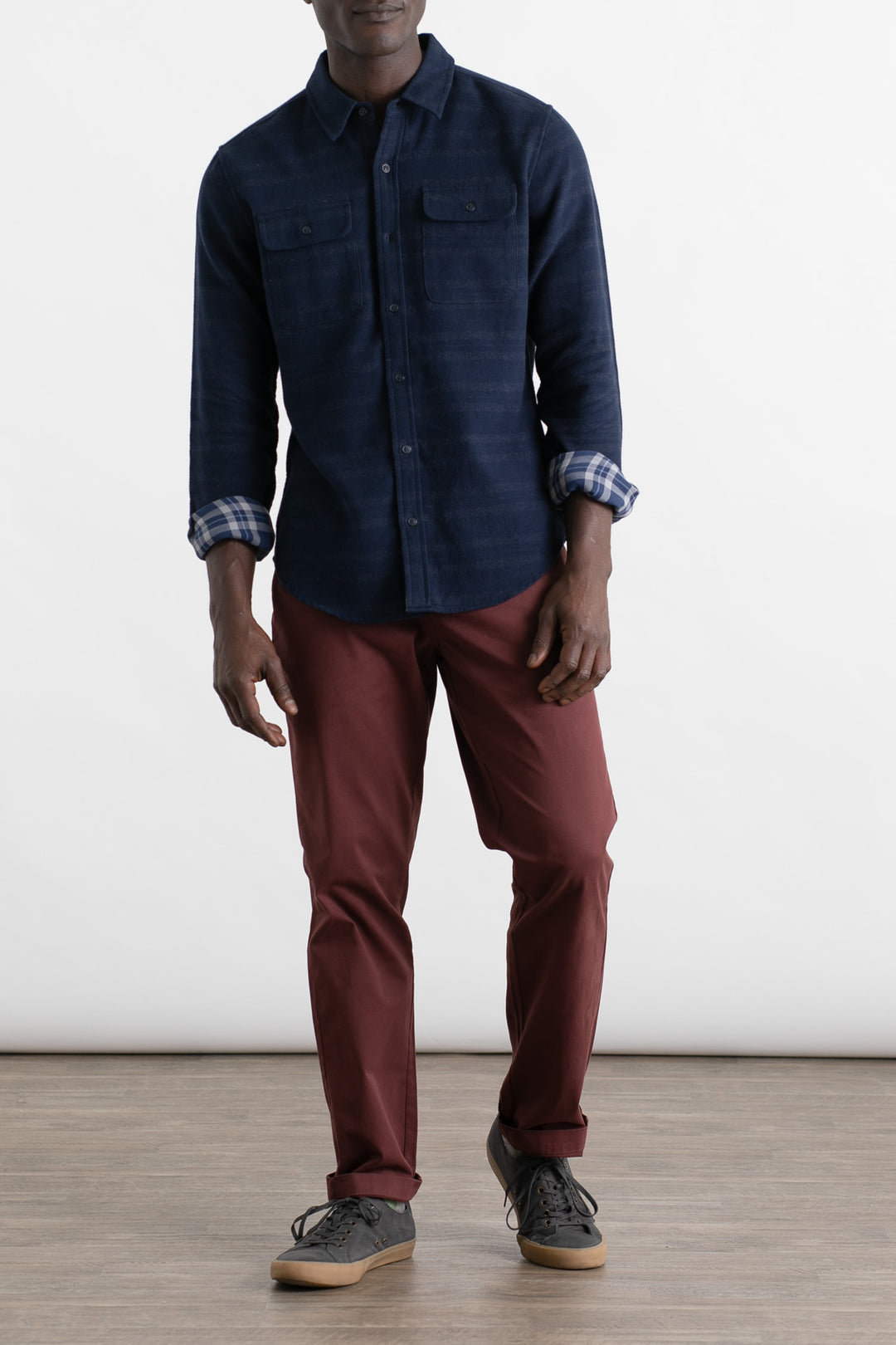 A person wearing a navy button-up shirt, Bridge & Burn Polk Pant in burgundy, and black sneakers stands on a wooden floor against a plain white background.