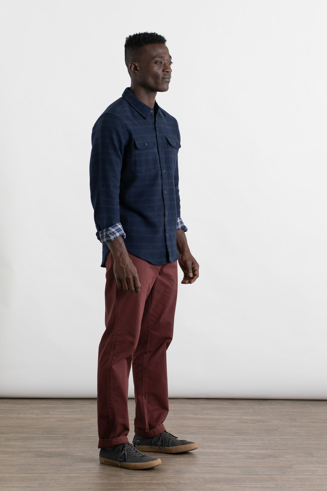 A man stands indoors against a plain white background, wearing a navy blue button-up, Bridge & Burn Polk Pant in burgundy, and gray sneakers. He faces slightly right and smiles gently.