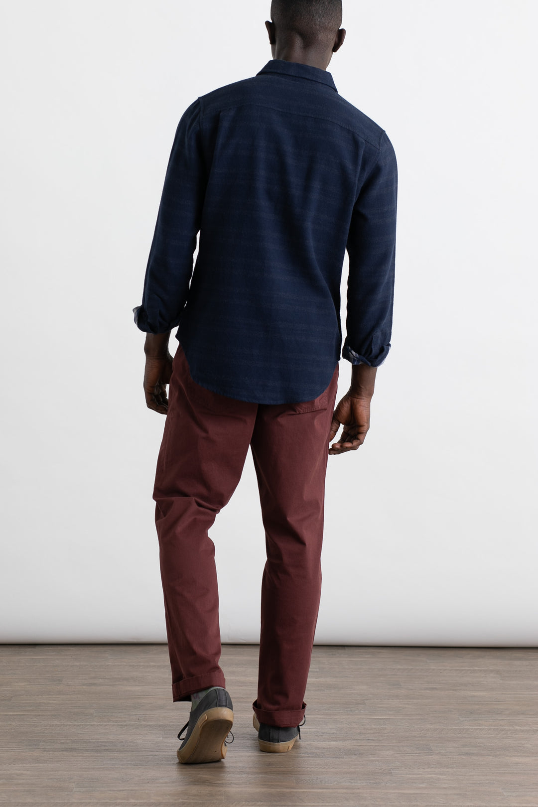 A person faces away from the camera, wearing Bridge & Burn's Polk Pant in Burgundy—a classic 5-pocket organic cotton style—with a dark blue long-sleeve shirt and gray shoes, standing on a wooden floor against a white background.