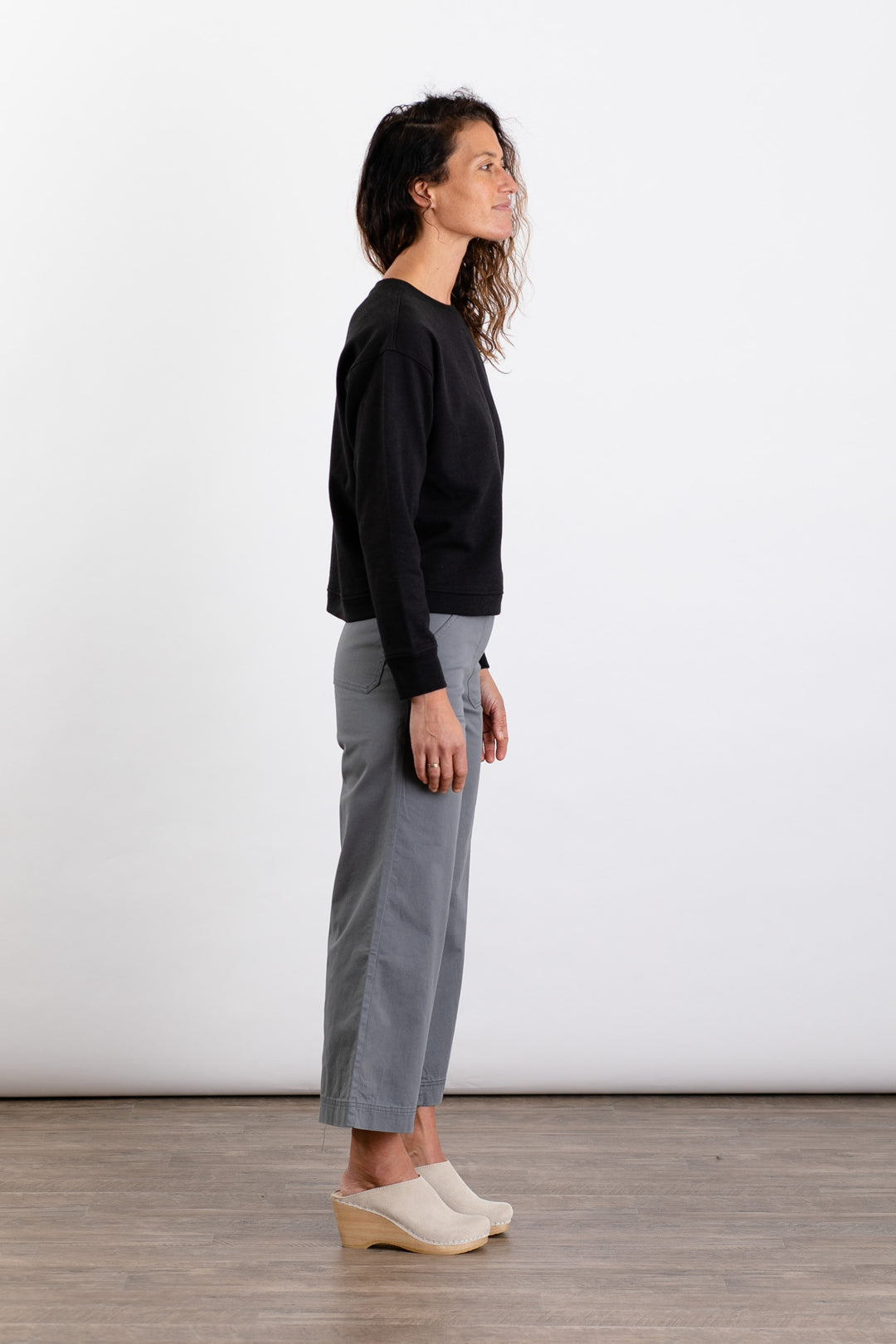 A woman with wavy hair stands in profile against a white background, wearing the Bridge & Burn Hawthorne Boxy Crew Sweatshirt in black, paired with gray cropped pants and light-colored clogs.