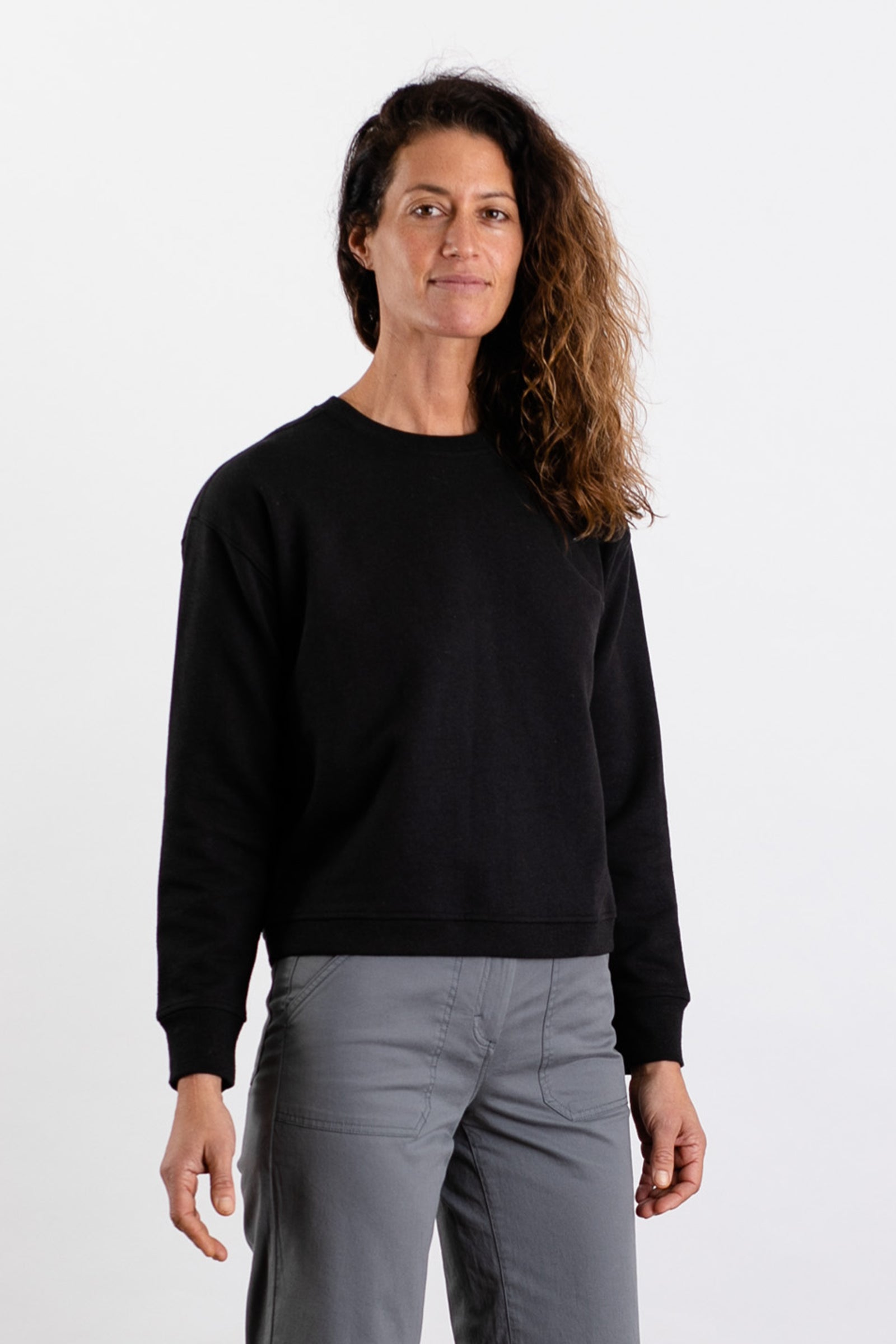 A person with long, wavy brown hair faces the camera, wearing the Bridge & Burn Hawthorne Boxy Crew Sweatshirt in black and gray pants against a plain white background.