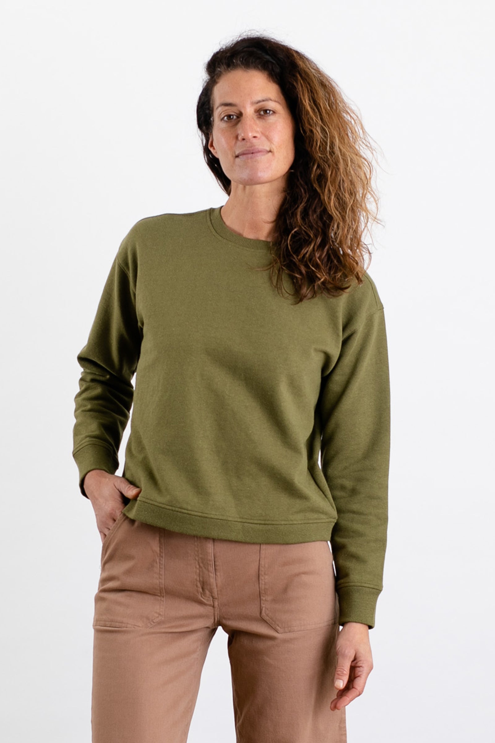A person with long, wavy brown hair stands against a plain white background wearing the Bridge & Burn Hawthorne Boxy Crew Sweatshirt in Olive and light brown pants, with one hand in their pocket and a neutral expression.
