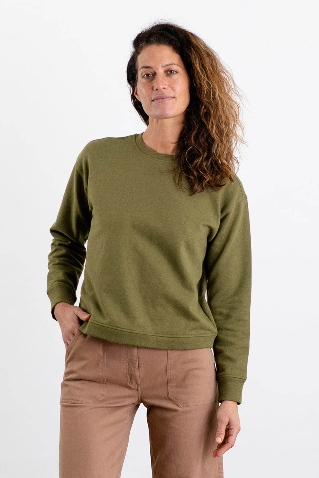 A person with long, wavy brown hair stands against a plain white background wearing the Bridge & Burn Hawthorne Boxy Crew Sweatshirt in Olive and light brown pants, with one hand in their pocket and a neutral expression.