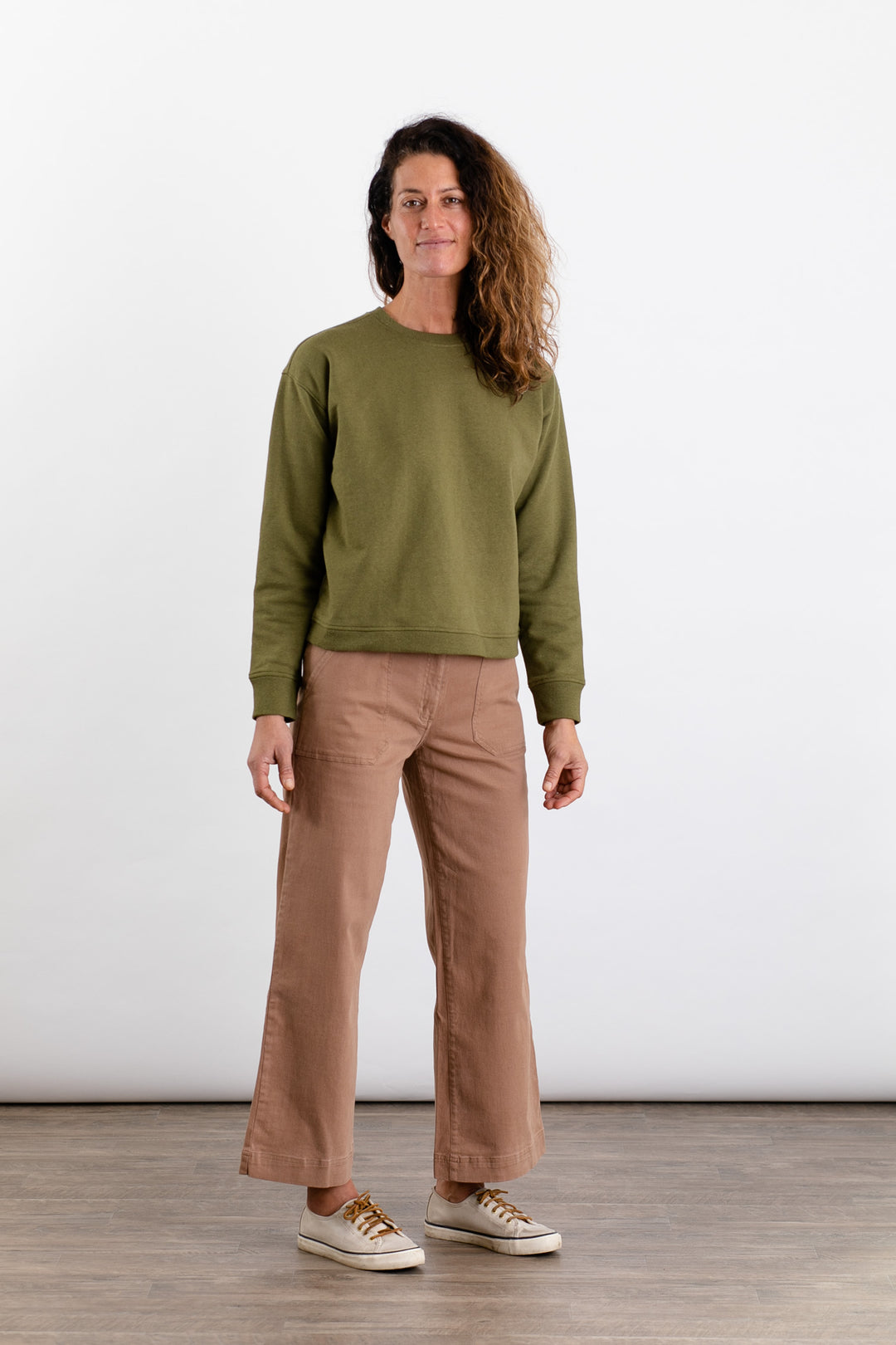 A woman with curly hair wears the Bridge & Burn Hawthorne Boxy Crew Sweatshirt in olive, light brown wide-leg pants, and white sneakers, standing on a wooden floor against a plain white background.