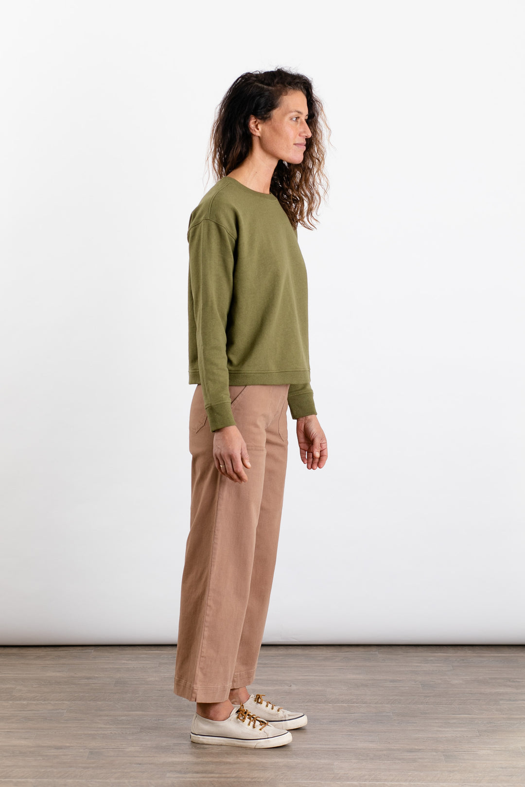 A woman with curly brown hair stands in profile on a wooden floor against a white background, wearing the Bridge & Burn Hawthorne Boxy Crew Sweatshirt in olive, tan wide-leg pants, and white sneakers.