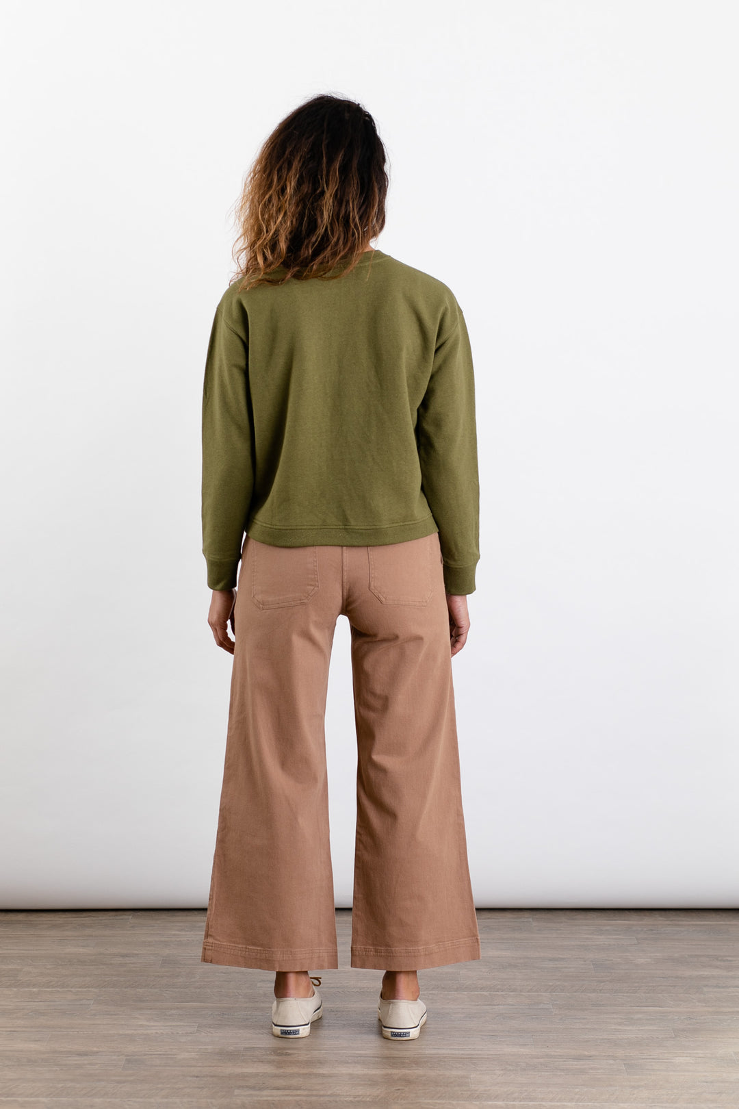 A person with wavy brown hair faces away, wearing the Bridge & Burn Hawthorne Boxy Crew Sweatshirt in olive, light brown wide-leg pants, and light-colored shoes against a plain white background.