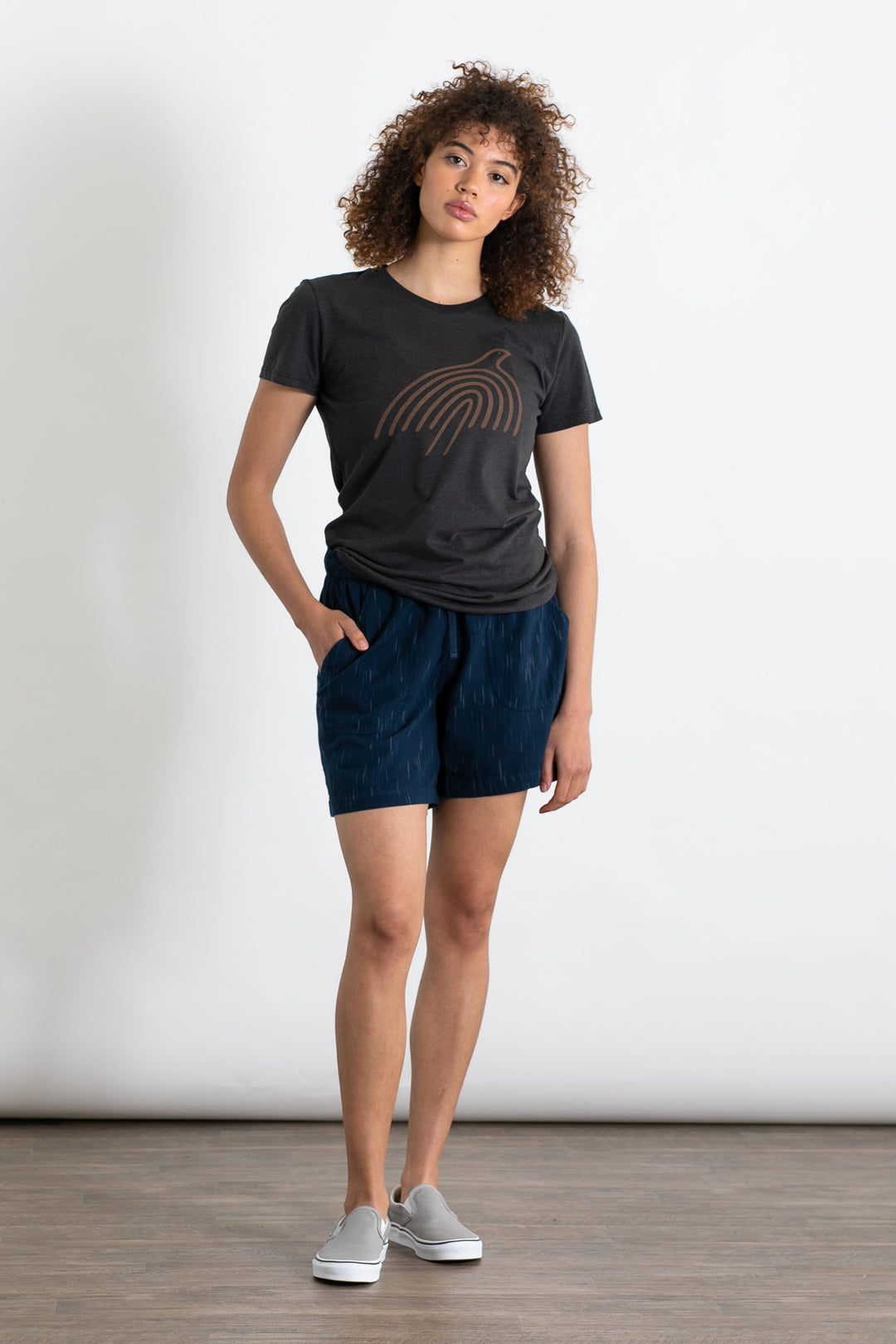 A person with curly hair wears a black graphic tee, Bridge & Burn’s Nico Short in Navy Space Dye with an elastic waist, and gray slip-on shoes, standing against a plain white background with one hand in their pocket.