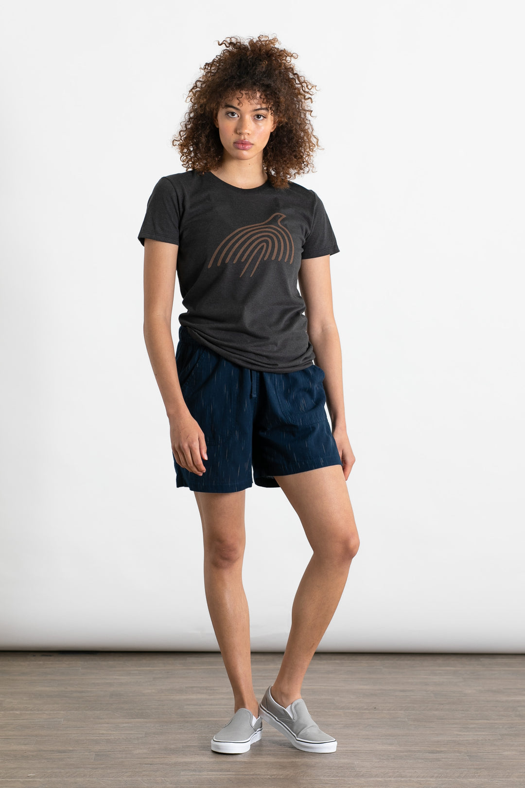 A person with curly hair wears a dark abstract tee, Bridge & Burn Nico Short in Navy Space Dye with an elastic waist, and gray slip-on shoes, standing against a plain white background.