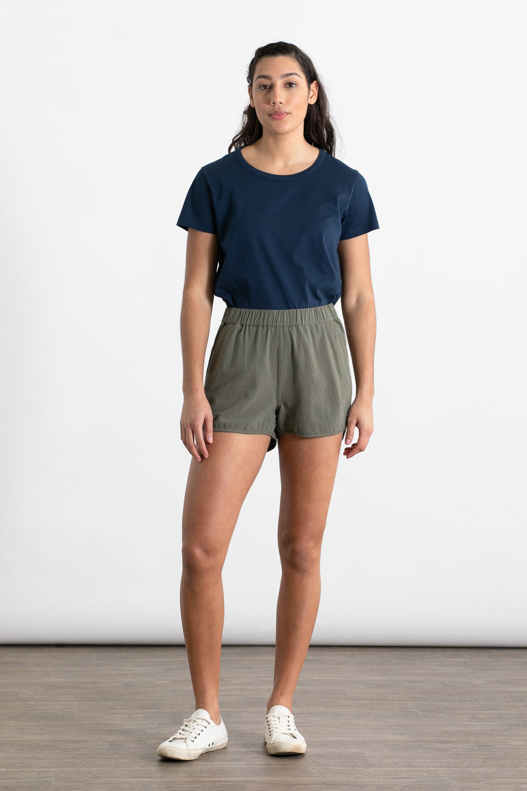 A person stands indoors against a white background, wearing a navy t-shirt, white sneakers, and Bridge & Burn’s Luca Track Short in Olive Pinstripe. They have long dark hair, a neutral expression, and their arms are relaxed at their sides.