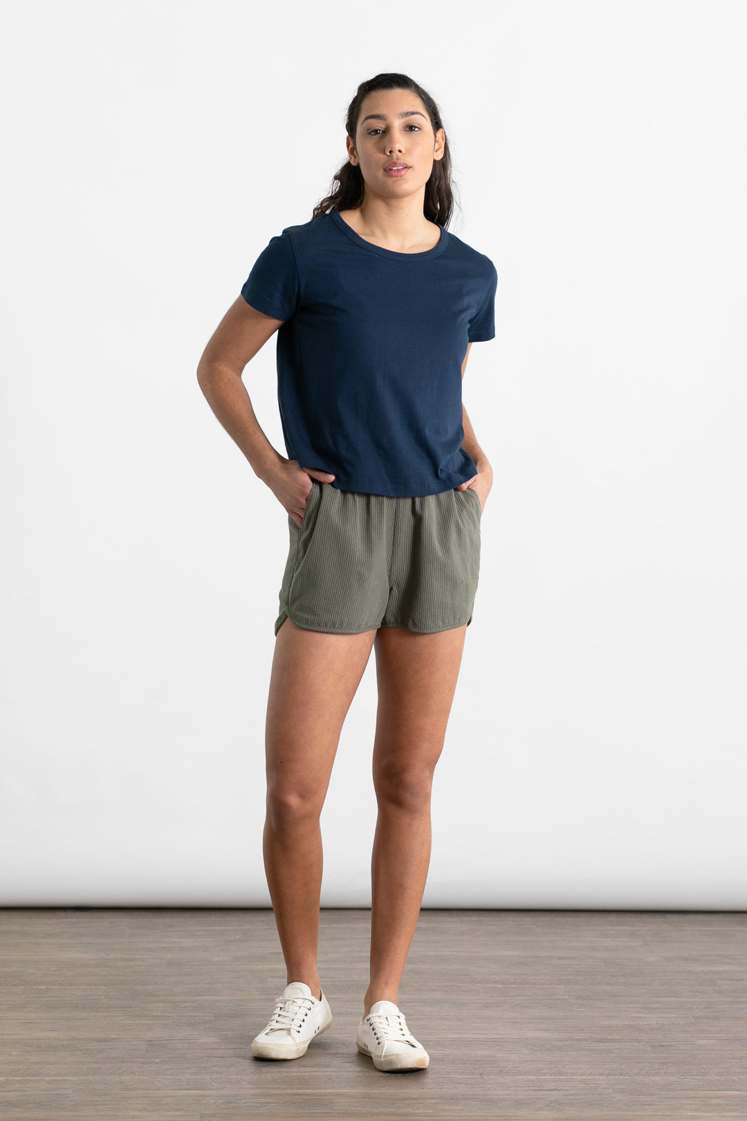 A woman stands indoors against a white background, wearing Bridge & Burn's Luca Track Short in Olive Pinstripe and a navy t-shirt. Her hands are in her pockets and she faces the camera with a neutral expression.