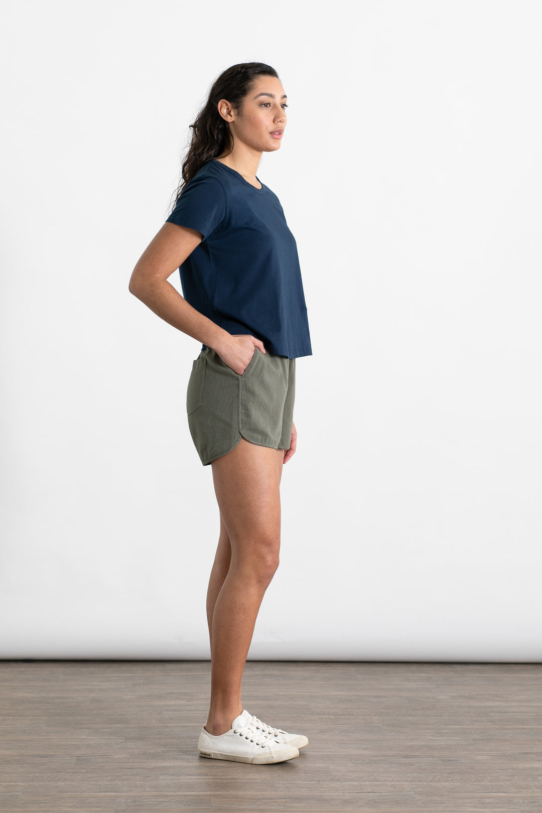 A woman stands in profile against a white background, wearing Bridge & Burn's Luca Track Short in Olive Pinstripe and a navy t-shirt. Her hands are in her pockets, and she has long, dark hair and white sneakers.