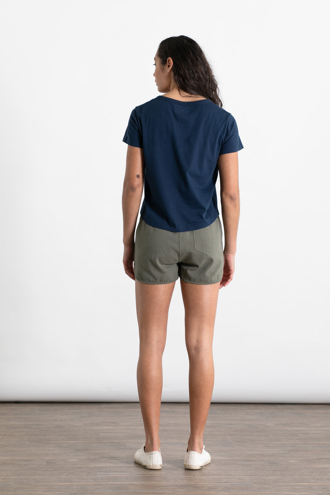 A person with long dark hair faces away, wearing a navy shirt, Bridge & Burn’s Luca Track Short in Olive Pinstripe, and white slip-on shoes, against a plain white background on a wooden floor.